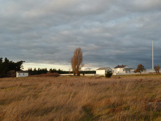 A collection of buildings, a picket fence, and a flagpole between a grassy field and low clouds.