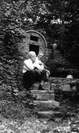 A man sits on a set of stone stairs, hand on his chin and facing away from the camera into trees