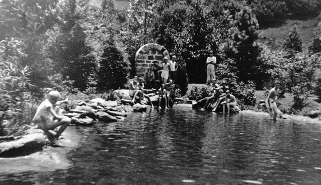 Bathers gather at the edge of a pool of water, near a brick structure marking a spring