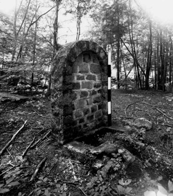 A rounded stone wall with a square basin on the ground, surrounded by woodland