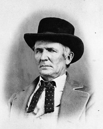 A portrait of a serious-looking man in a dark, brimmed hat and polka dot tie