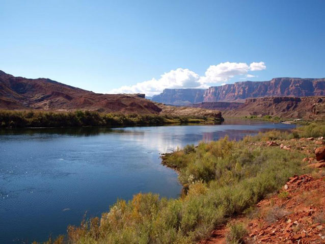 A wide blue river spreads through a canyon landscape, bordered by low scrub and rock walls