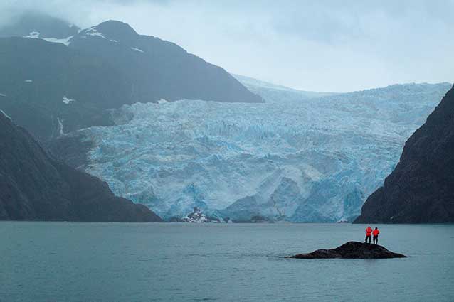 two people standing on a tiny rock island in a fjord, looking at a massive tidewater glacier