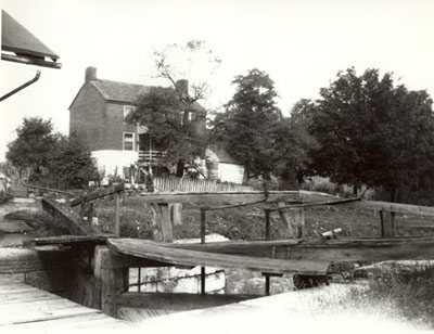 Rough wooden walkway passes over a narrow waterway towards a two-story farmhouse, with trees. Rough wooden walkway passes over a narrow waterway towards a two-story farmhouse, with trees.