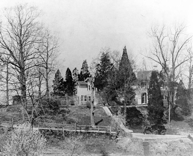 A horse-drawn wagon on a road below a house atop a hill, surrounded by trees A horse-drawn wagon on a road below a house atop a hill, surrounded by trees