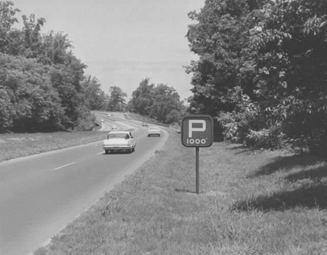 Historic image of a car driving on a parkway, bordered by a grassy shoulder and trees Historic image of a car driving on a parkway, bordered by a grassy shoulder and trees