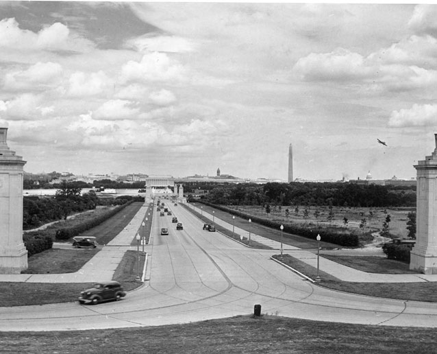 Cars cross a bridge toward Lincoln Memorial on a wide road, bordered by landscaping and lampposts