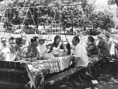 A crowd gathers on both sides of a picnic table, covered with food and a tablecloth