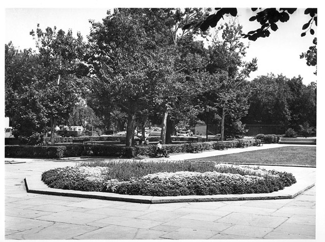 A low-growing flower bed in a central stone plaza with trees, hedges, and lawn beyond.