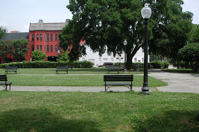 Buildings stand beyond the park perimeter, viewed across an area with benches, grass, and walkways.
