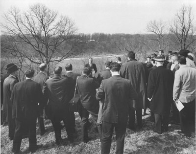 A man speaks to a crowd in suits and overcoats, all standing in front of a wide view. A man speaks to a crowd in suits and overcoats, all standing in front of a wide view.