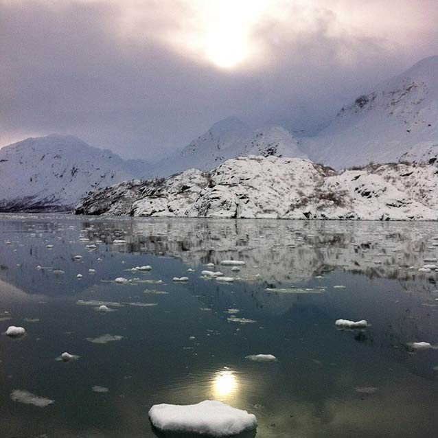 sun setting behind thick clouds, over snowy mountains bordering an ice-filled bay