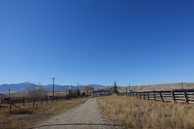 A farmhouse at the end of a straight dirt drive, framed by dry grass, wooden fences, and blue sky.