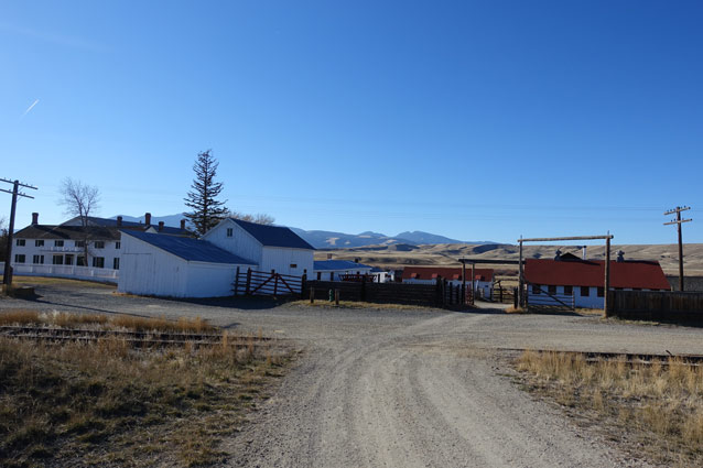 A gravel driveway crosses railroad tracks towards a cluster of farm buildings and fencing.