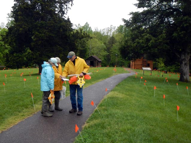 Three people in raingear on a paved path, surrounded by survey markers in the grass