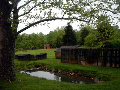 The cabin and survey flags are surrounded by a small pond, wooden fence, and large trees