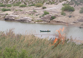 A firefighter in a canoe on the river observes fire on the riverbank.