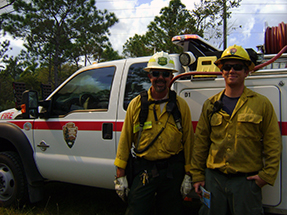 Two firefighters stand near a wildland fire engine. Two firefighters stand near a wildland fire engine.