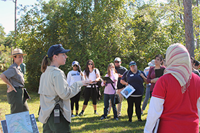 A group stands in a half circle listening to a woman speaking to them.