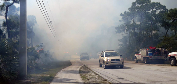 Vehicles drive on smoky roadway with fire vehicles parked on roadside.