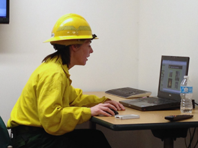 A firefighter in Nomex flame-resistant gear sits in front of a computer at a desk.