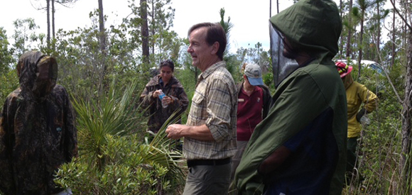 Three people stand talking in an open area near vegetation and pine trees.