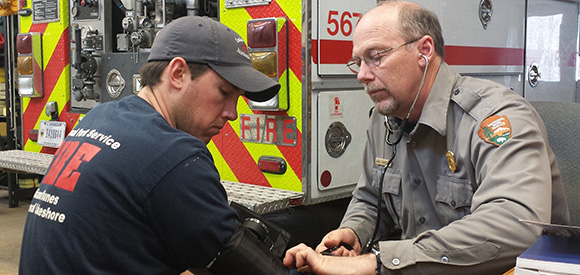 A man in a National Park Service uniform uses a blood pressure cuff on another man. A man in a National Park Service uniform uses a blood pressure cuff on another man.