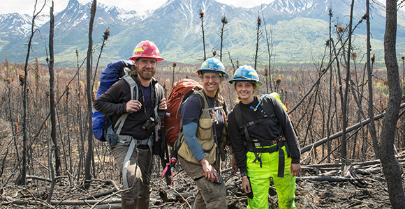 Two men and one woman, all in hardhats, stand in a burned area.