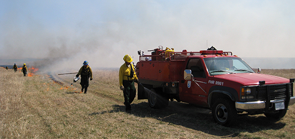 A firefighter walks near a wildland fire engine while a fire burns in grass in the distance.