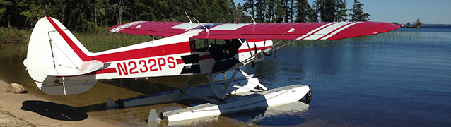 A fixed wing airplane of floats sits on the shore of a lake.