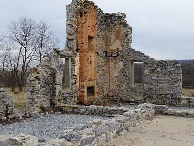 A corner of a stone wall with a brick chimney and windows stands in an open field.