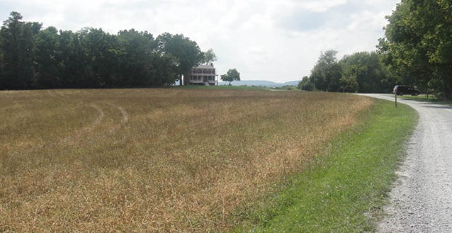 A driveway curves past flat fields and wooded areas towards a large farmhouse.
