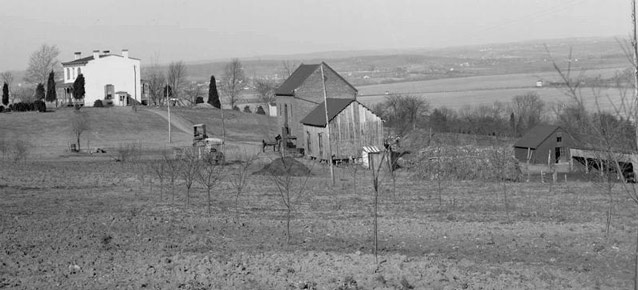 A distant view of a two-story farm house surrounded by fields, farm buildings, and expansive views.