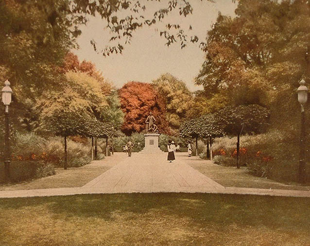 A wide, straight walkway between symmetrical ornamental plantings leads towards a statue