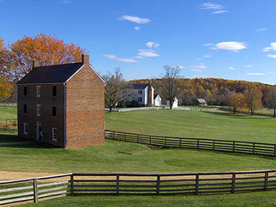 A rural landscape with a three-story brick buildings, house in the background, and wooden fencing.