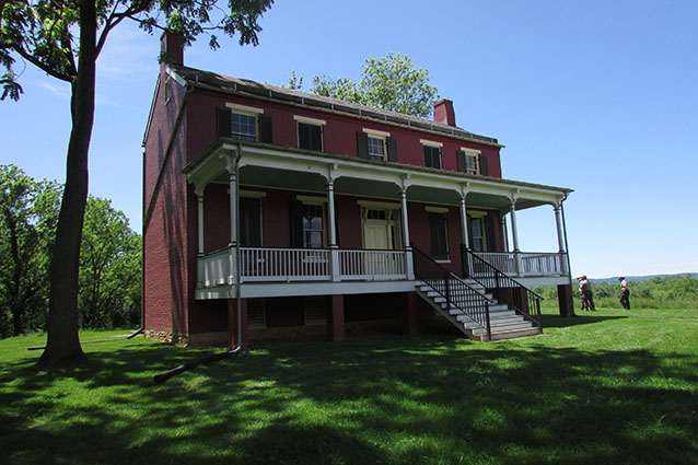 A two-story red farmhouse has a porch across its front length with stairs in the center.