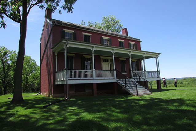 A two-story red farmhouse has a porch across its front length with stairs in the center. A two-story red farmhouse has a porch across its front length with stairs in the center.