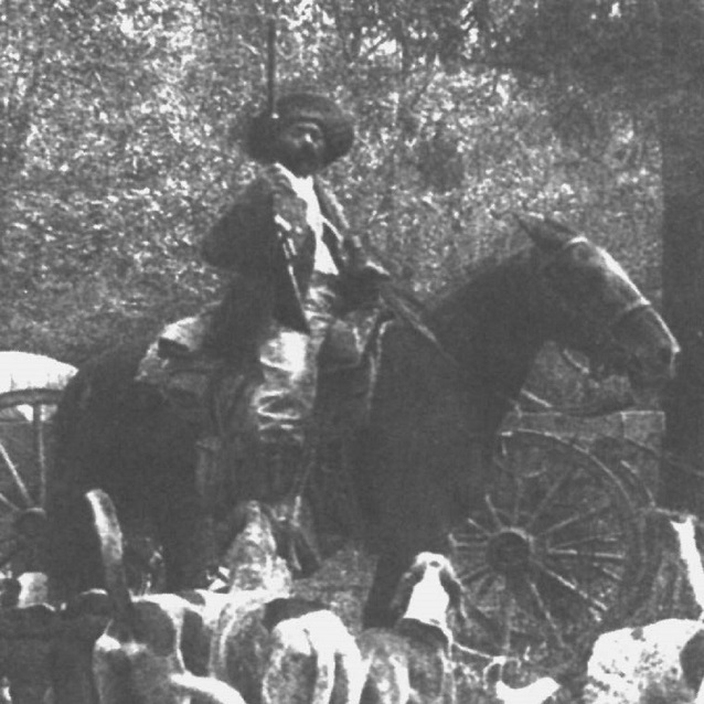 black and white image of man on horse, with gun, and hounds at his feet. 