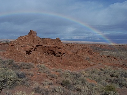 A red sandstone pueblo with shrub vegetation against a blue with wispy clouds and a full rainbow.