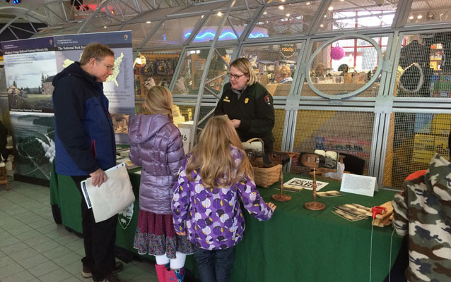 Adult and two kids talking to a park ranger at a table. Table covered with items