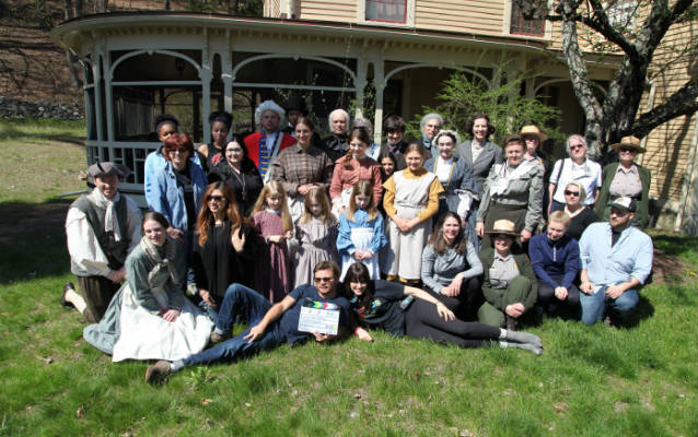 Group of people, some in historic clothes, many in modern clothes, pose for a photo outside The Wayside, in Concord, MA.