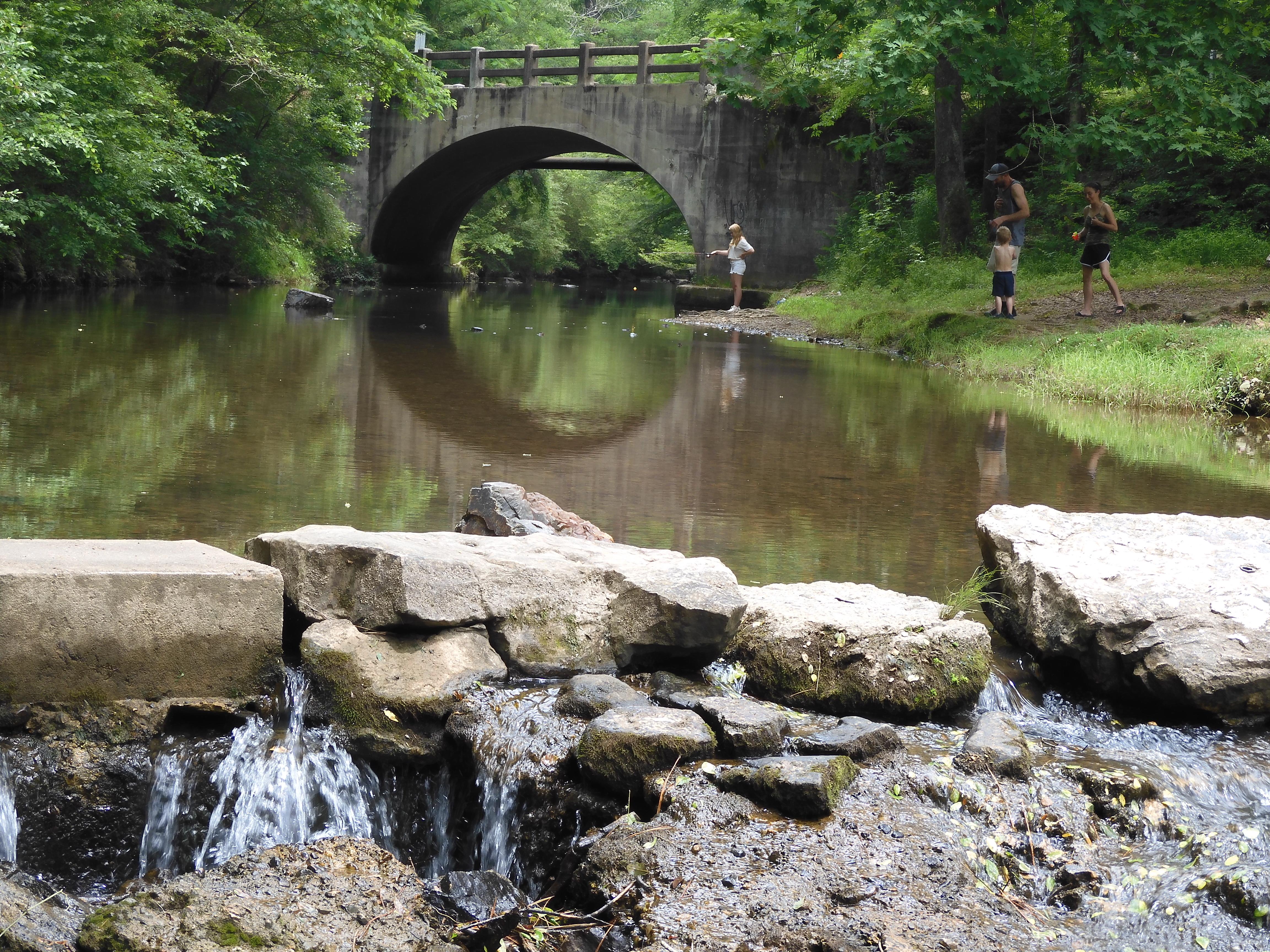 Two people stand on the banks of Gulpha Creek. An arched bridge is reflected in the water.
