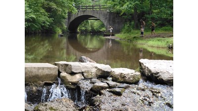 Two people stand on the banks of Gulpha Creek. An arched bridge is reflected in the water.