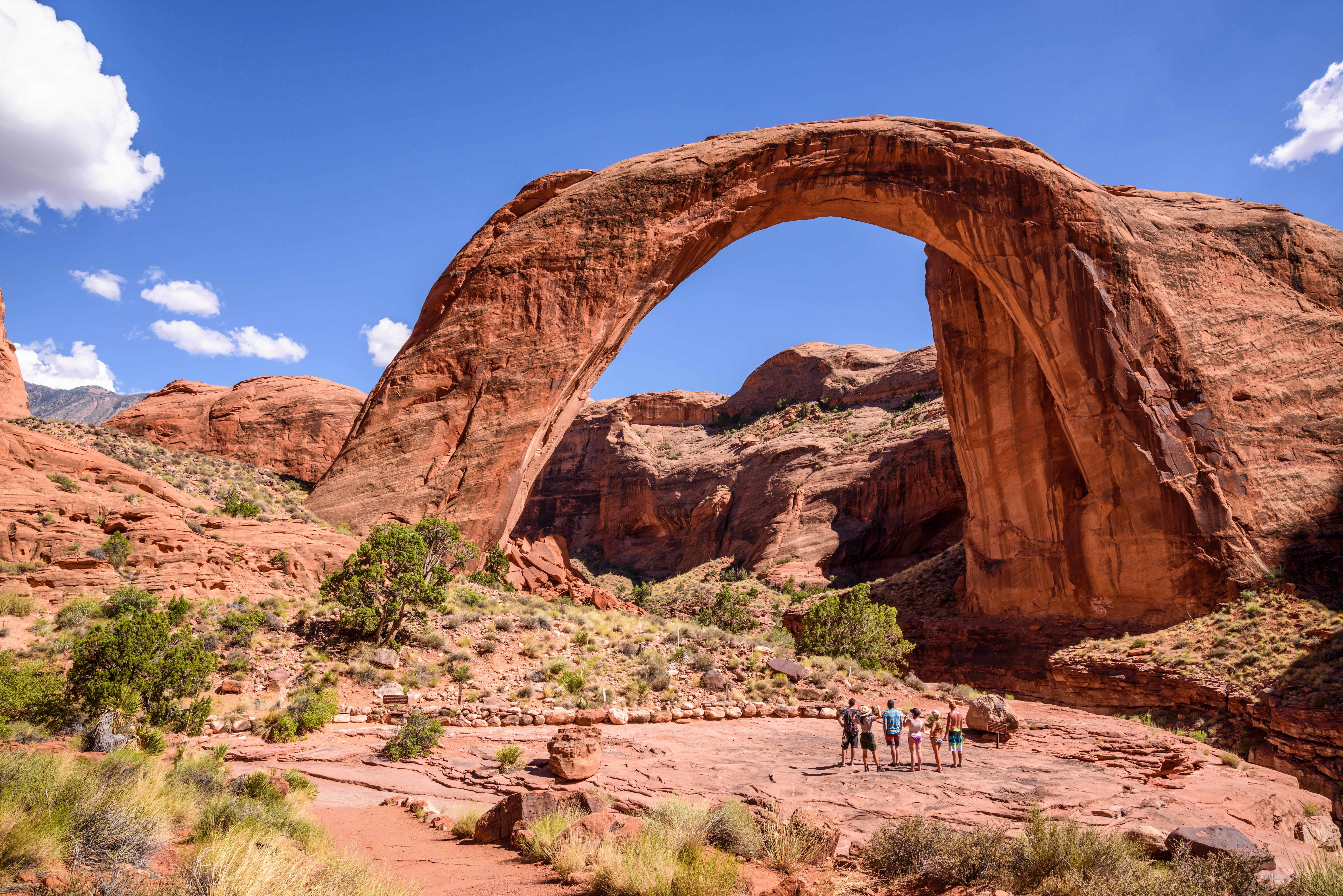 A large natural rock arch spans a canyon with people looking up at it.