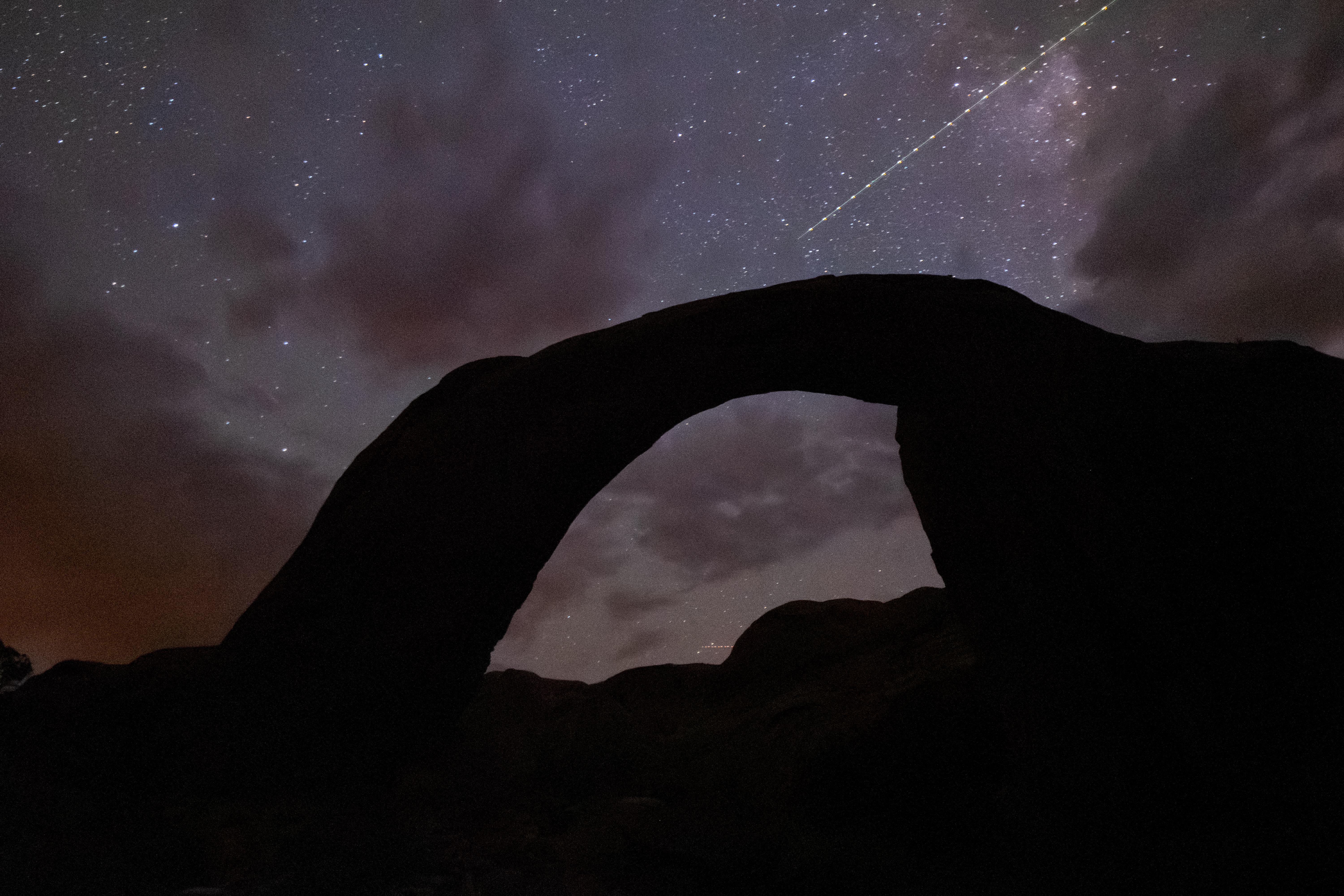 A silhouetted rock arch under a stary sky with a satellite flying by.