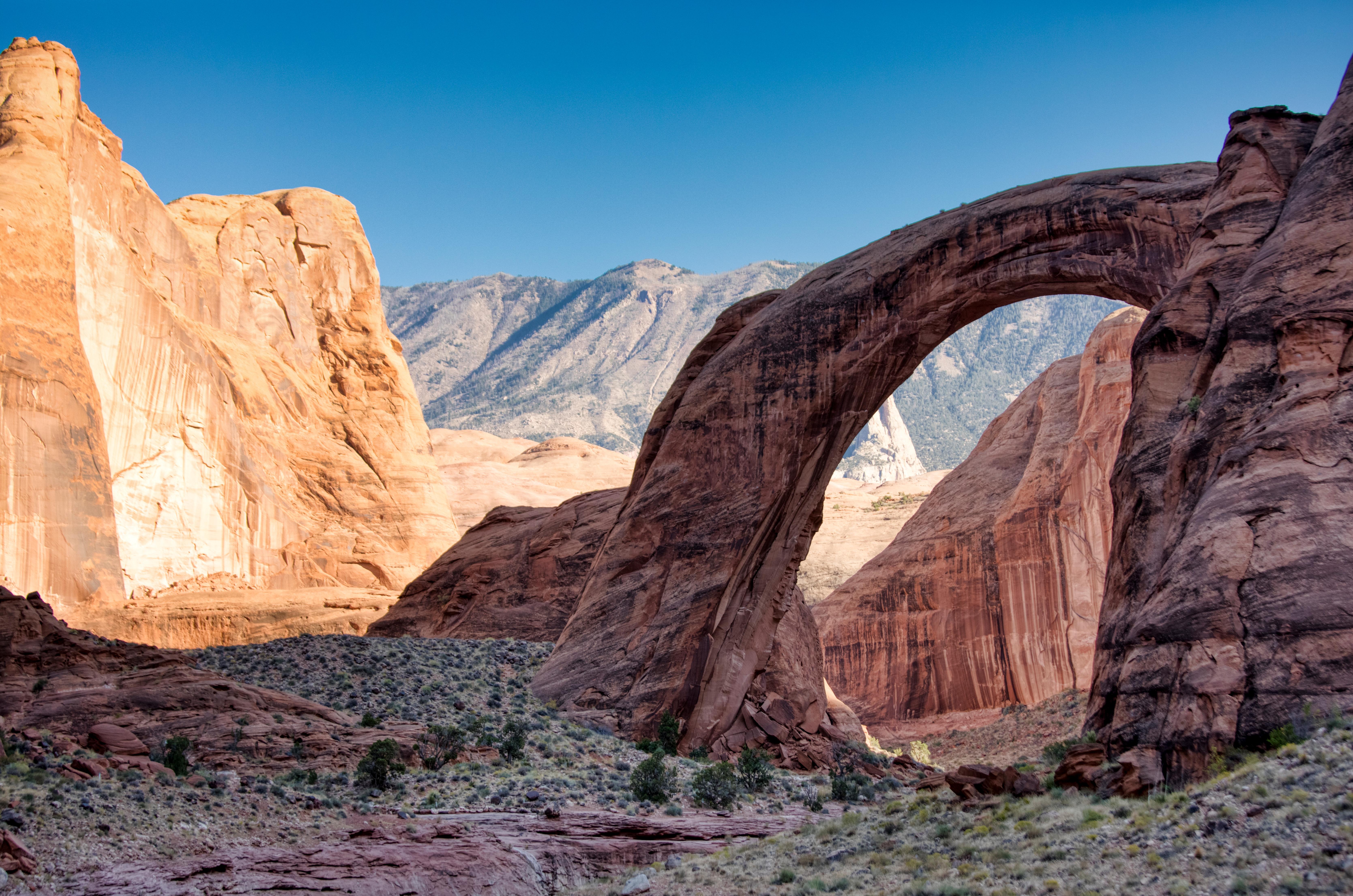 A natural stone arch sits between red rock canyon walls.  A mountain is seen through the arch.