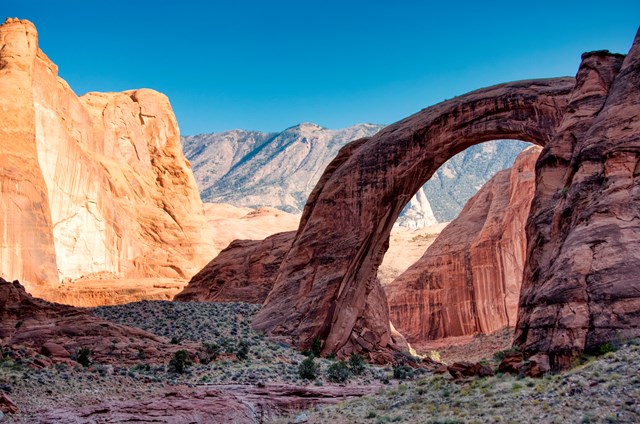 A natural stone arch sits between red rock canyon walls. A mountain is seen through the arch.