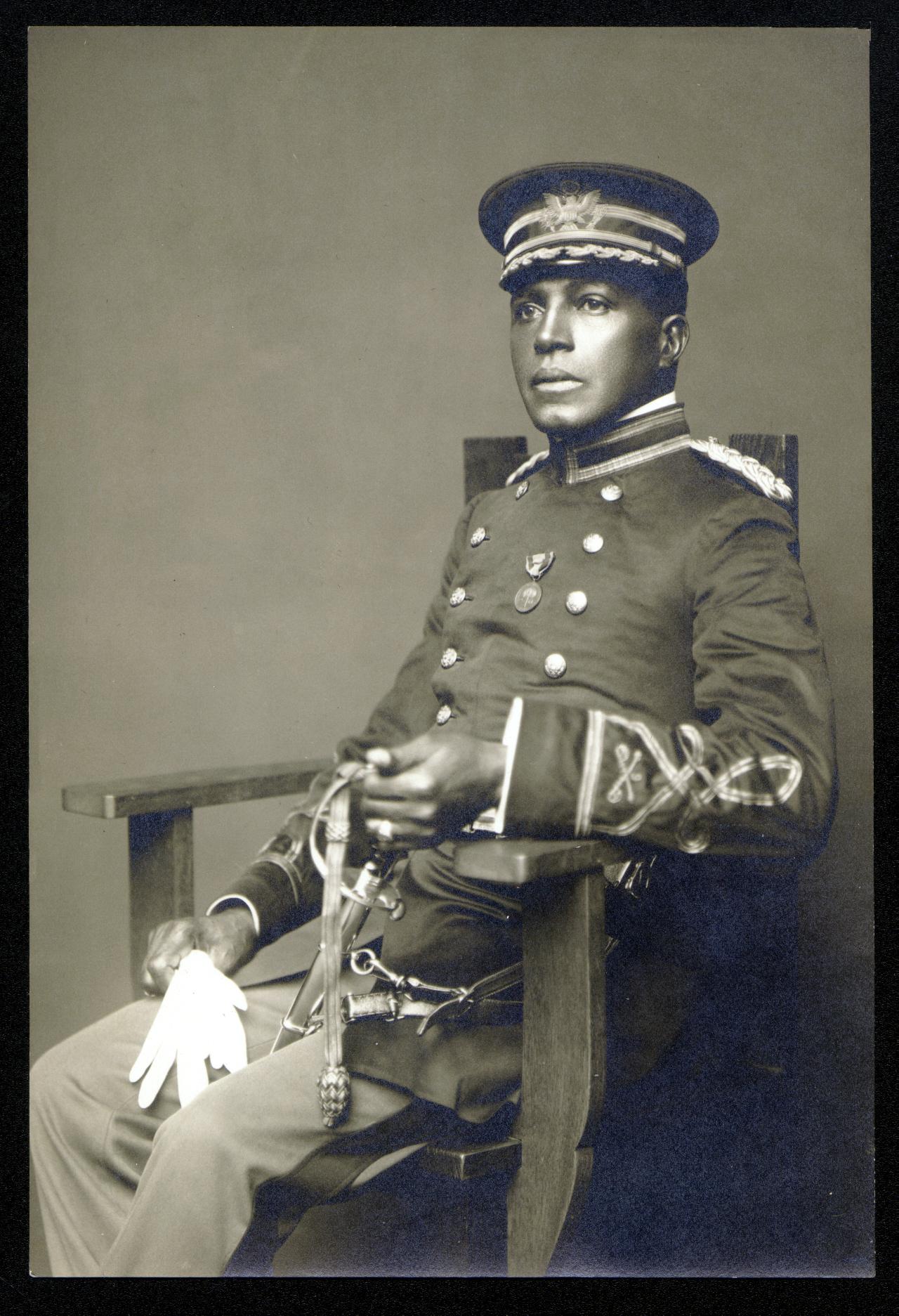 An African American man in a military uniform holding a sabre and wearing a cap sitting in a chair