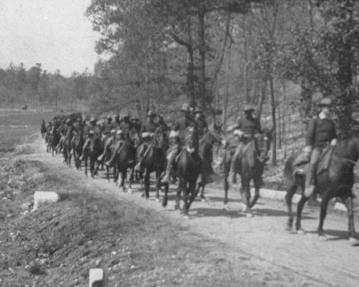 Several african american soldiers on horseback parading down a dirt road.