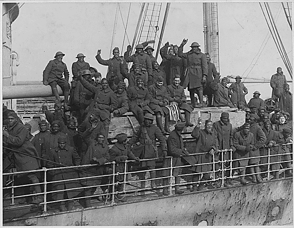 Several African American soldiers waving while standing and sitting on a boat's deck